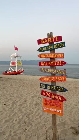 A wooden signpost with multiple directional arrows stands on a sandy beach. The arrows are painted in different colors and point in various directions, each indicating a beach name such as Sarti, Achlada Beach, Kavourtrypes, Kalamitsi, Platanitsi Beach, Paradise Beach, Akti Klimataria, and Goa Beach. The last arrow indicates Sarti Hotel Association with an Instagram handle. In the background, a lifeguard tower with a red flag is visible, along with a red kayak on the sand. The sea stretches out behind the beach, and distant hills can be seen on the horizon. The sky is a pale blue, suggesting it is either early morning or late afternoon. The sand is light brown and appears undisturbed.
