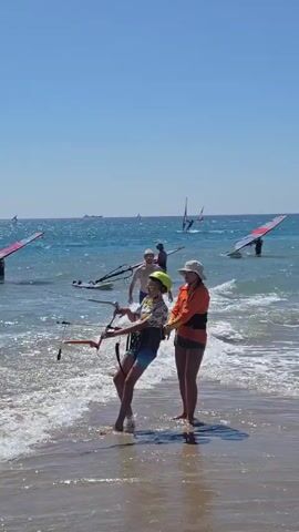 A child wearing a yellow helmet and a multi-colored shirt stands in shallow water at the edge of a sandy beach, holding onto a windsurfing boom. An adult in an orange vest and a white hat stands behind the child, placing their hands on the child's back and the boom, appearing to assist or instruct. In the background, several other individuals are windsurfing on the blue ocean under a clear sky. The waves are breaking on the shore.
