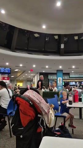 The video shows a busy indoor space, likely an airport or mall food court.  People are seated at tables and walking around. In the foreground, a stroller is visible.  Several individuals are sitting at tables, some with drinks and laptops.  One child wearing headphones is at a table.  In the background, there are shops and signs, including one that says "HOUSE OF FRIENDS".  The ceiling features a large, circular design with screens or panels. The lighting appears to be artificial.