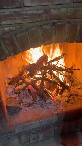 The video shows a close-up of a brick fireplace with a roaring fire. The fire is built with logs and branches, with flames reaching high within the confines of the hearth.  Ash and embers are visible beneath the burning wood. Music can be heard in the background.