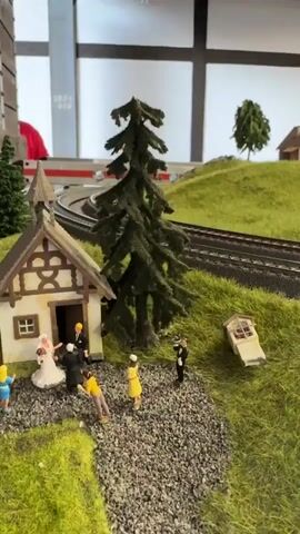The video shows a miniature model train set with a wedding scene in the foreground. A small, quaint house with a thatched roof sits to the left, with several miniature figures gathered outside. A bride in a white gown and a groom in a dark suit are among the figures, suggesting a wedding ceremony is taking place or has just concluded. Other figures, including a person in a yellow dress and a person in a black suit, are also present. To the right of the house, a small structure resembling a shed or a chapel lies on its side, as if it has fallen over. The train tracks, which are elevated on a green, grassy landscape, curve around the scene and continue into the background. A miniature evergreen tree stands tall near the house, and other trees are scattered across the landscape. In the background, a train is seen passing by on the tracks, adding movement to the otherwise static scene. The environment appears to be an indoor space with industrial-style lighting and a ceiling with exposed beams.