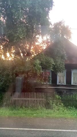 The video shows a wooden house with two windows and a fence in front of it. The house is surrounded by trees and bushes. The sky is lit by the setting sun, casting a warm glow over the scene. The video appears to be shot from a moving vehicle, as evidenced by the slight blur and the road in the foreground. The scene is peaceful and idyllic, evoking a sense of rural tranquility.