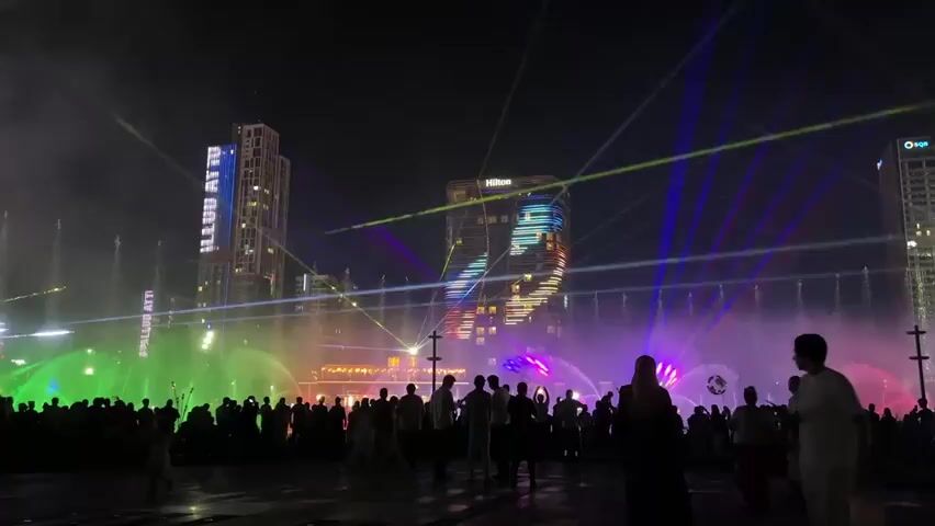 The video captures a nighttime scene in a city plaza where a large crowd of people are gathered to watch a water and light show. Tall buildings, illuminated with colorful lights and laser beams, form the backdrop. One building prominently displays a light show with moving patterns. In the foreground, a fountain shoots jets of water that are lit with vibrant green and pink lights, creating a misty atmosphere. People of various ages, dressed in casual clothing, are visible in silhouette against the bright lights, some walking and others standing to observe the spectacle. Several individuals are pointing or looking towards the main attraction. The ground appears wet, suggesting the mist from the fountains has settled. The overall ambiance is one of celebration and public entertainment, with the dynamic interplay of light, water, and architecture creating a visually striking display for the assembled audience.