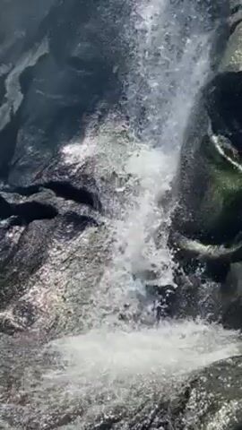 The video shows a waterfall cascading over dark rocks. Splashes and mist rise from the bottom of the falls. Someone shouts joyfully, but they are not visible.