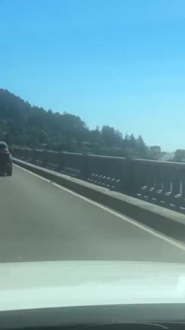 A black SUV drives down a two-lane road. The road has yellow lines separating the lanes. There is a concrete barrier to the right of the road with arched openings. Behind the barrier, there is water visible. The road is on a bridge, as indicated by the barrier. There is a green sign on a pole to the right of the road. The sign says "Jerrys Flat Rd" and "LEFT 1/4 MILE". Trees and houses are visible on the hillside to the left of the road, under a bright blue sky. The image is taken from inside a vehicle, as indicated by the blurry white area at the bottom of the frame.