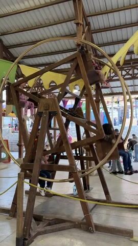 The video shows a wooden Ferris wheel structure indoors, with several children riding in it. The wheel is made of natural-colored wood, constructed in a rustic or medieval style. Children are seated in open-air compartments built into the wheel's frame. The wheel is being manually rotated, likely by an operator not visible in the frame, causing the children to go up and down. 

The setting seems to be a fair, festival, or market, with people walking around in the background. The floor is light-colored concrete, and there are various decorations and stalls visible in the background, including yellow and green fabrics. Overhead, the building has an open, exposed rafter ceiling. A water bottle is visible on the ground near one of the wheel's supporting legs. People in the background are dressed in everyday clothing.