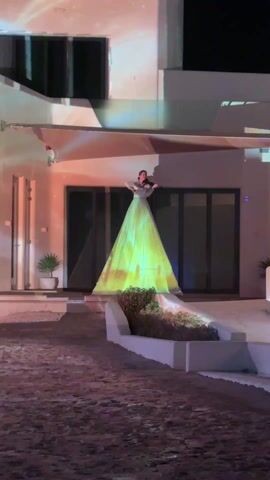 The video shows a woman standing on a slightly elevated patio in front of a modern, white house at night. She is playing a violin. She is wearing a light-colored top and a floor-length gown that appears to be white but has a dynamic, light-based projection on it that includes bright yellow and swirling light effects. The projection also seems to extend onto parts of the house behind her, creating a blended visual effect. The patio is white, surrounded by a bed of green plants framed by white concrete. Below the patio, the ground is covered with a reddish-brown sand or dirt. The house has sliding glass doors behind her, and there are small potted plants near the entrance on the left.