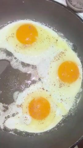 The video shows a close-up, overhead shot of three eggs being fried in a black skillet. The eggs are sunny-side up, with bright orange yolks and cooked whites. They are seasoned with black pepper. The oil in the pan is bubbling around the eggs, indicating they are actively cooking. The skillet appears to be cast iron.