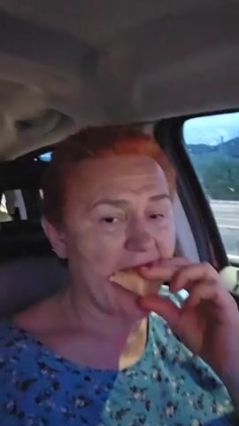 A woman with short red hair is sitting in the driver's seat of a car and eating a croissant. She is wearing a blue floral print shirt. The video is shot from a close-up angle, focusing on her face and the croissant. The background is blurred, but it appears to be dark outside, possibly nighttime or early morning, with some lights visible in the distance. The car interior is dimly lit.