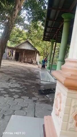 A person wearing a blue shirt and a pink headscarf is sweeping the ground in front of a small wooden house. The house appears to be in disrepair, with clothes hanging on a line outside. The ground is paved with stones, and there is a tree to the left of the frame. In the foreground, there is a raised structure with decorative carvings. The scene suggests a rural or semi-rural setting.