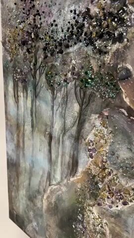 The video shows a close-up, panning view of a mixed-media artwork. The artwork depicts a serene female figure with closed eyes, seemingly in repose. The figure's face is rendered with subtle shading and muted colors. Her hair and parts of her body are adorned with a dense application of various materials, including black and dark-colored stones or beads, shimmering purple gemstones, and iridescent glitter. These elements create a textured, almost jewel-encrusted effect. In the background, abstract representations of trees with purple accents are visible, rendered in a soft, painterly style. The overall impression is one of mystical beauty and ethereal tranquility.
