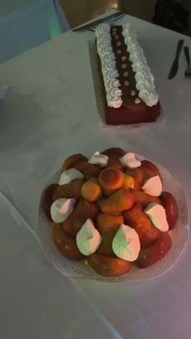 The video begins with a close-up of a round plate filled with what appear to be small, glazed pastries topped with dollops of whipped cream and some round, orange-colored fruits. In the background, slightly out of focus, is a rectangular cake decorated with white frosting and brown chocolate accents. The scene is set on a white tablecloth, possibly in a dimly lit room, with some cutlery visible in the background.

The camera then pans to the right, showing a rectangular cake with swirled frosting in yellow and white, and a fork resting beside it. A lit candle in a glass holder is prominent in the foreground, casting a warm glow.

Further panning reveals more of the table setting. There are stacks of white plates, a small wooden-topped stand with metal legs, and another rectangular cake in the background, this one topped with green and yellow frosting and chocolate drizzle. There's also a tray of what looks like donuts or small round pastries further down the table. The lighting suggests an evening event, possibly a party or celebration. The video ends with a view of the lit candles again.