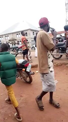 The video shows an outdoor scene in what appears to be a marketplace or busy street. The ground is red dirt. Several motorcycles are visible. In the foreground, a person wearing a tan shirt, white shorts, and sandals walks away from the camera. A child in a green vest and yellow pants walks towards the camera in the lower left corner. In the background, a group of people are gathered. One person, a young male wearing an orange shirt with black stripes, stands facing left, holding something to their ear, possibly a phone. A woman in a colorful dress is also in the group. A building with a light-colored facade and multiple windows is behind the group. The sky is overcast.