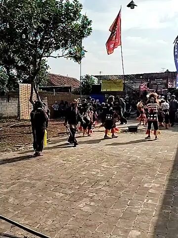 This video captures an outdoor cultural performance in Mojodoyong, Indonesia. The scene features a group of performers in traditional attire, characterized by elaborate headdresses and colorful costumes, engaged in what appears to be a lively dance. Several individuals are dressed in black, one holding a yellow sash, potentially indicating a directorial or ceremonial role. They are performing on a paved area, likely a village street or square, with spectators gathered on the sides.

In the background, traditional Indonesian houses with tiled roofs are visible, along with trees and fencing. A bright red flag flies prominently from a pole, adding to the festive atmosphere. Banners with text are also present, though the specific content is illegible due to the video's resolution. The weather appears to be clear and sunny, suggesting it is daytime. The overall mood seems to be celebratory and communal, highlighting a cultural event or festival. The presence of a modern light fixture overhead hints at the event potentially extending into the evening or being held in an area that uses electricity. The focus is on the dynamic movements of the performers and the engagement of the audience.