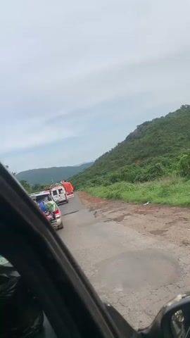 The video shows a view from inside a vehicle of a road with several vehicles in front. The road appears to be in a rural area, with green hills on the right side of the frame. The road surface is cracked and uneven. In front of the vehicle, there is a line of vehicles including a white van or truck, a white vehicle that might be an ambulance, and a light-colored vehicle loaded with items. There are also two people visible in orange shirts standing on the side of the road near the front vehicles. The sky is cloudy.