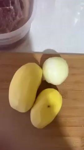 The video shows ingredients for a meal preparation. A clear plastic container holds raw meat submerged in water. On a wooden cutting board, there are two peeled potatoes and a peeled onion. The environment appears to be a kitchen.