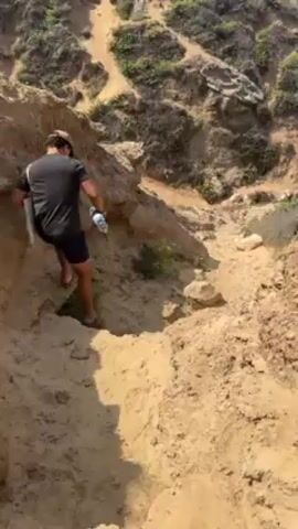 A man in a black t-shirt, shorts, and a cap walks barefoot down a steep, sandy cliff path. He's carrying a reusable bag and a water bottle. The path is narrow and rocky, with vegetation on the sides. The ocean and beach are visible in the background.