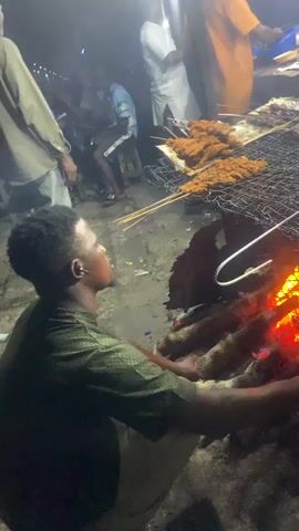 A man in a green shirt kneels in the foreground, tending to a fire. In the background, several people are gathered around a grill where food is being cooked over coals. The scene appears to be outdoors at night, with artificial lighting illuminating the area.