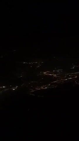 The video shows an aerial view of a city at night. The city lights are visible as a grid of yellow and white dots against the dark landscape. There are some faint streaks of light, likely from moving vehicles. The overall impression is one of a vast, illuminated urban area stretching into the distance.