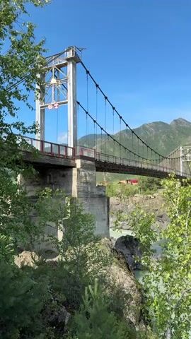 The video shows a suspension bridge crossing a turquoise river. The bridge has two large white towers that support the suspension cables. The bridge deck is red. The surrounding environment is mountainous with green hills. There are trees on the left side of the bridge, and rocks and vegetation in the foreground. The sky is clear and blue. The water in the river appears to be moving swiftly.