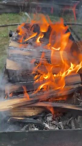 A fire is burning in a charcoal grill. The grill is rectangular and made of metal, with a pile of split firewood inside. The wood is burning vigorously with bright orange flames and producing smoke. The grill is situated outdoors on a grassy lawn. A wooden fence is visible in the background.
