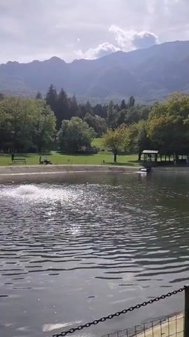 The video shows a serene lake with a fountain spraying water in the foreground. In the background, there is a grassy area with trees, some people, and a small gazebo. Beyond the trees, majestic mountains rise against a cloudy sky. The overall atmosphere is peaceful and natural.