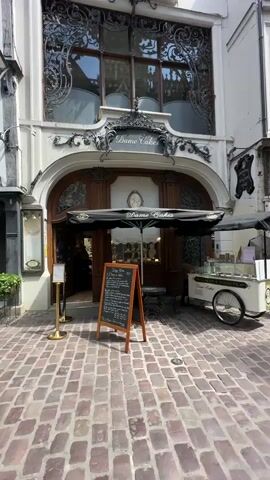 The video shows the exterior of a building with the signage "Dame Cakes". The building has an ornate facade with large windows featuring decorative metalwork. There is an arched entrance with a black awning that also says "Dame Cakes". In front of the entrance, there is a chalk board menu on an easel and a small table with two chairs. To the right of the entrance, there is a white cart with a black umbrella and signage that reads "Dame Cakes" and "Glace et Sorbets". The ground in front of the building is paved with red bricks. The sky is visible at the top of the frame. The video pans upwards slightly, focusing on the architecture of the building. No people are clearly visible interacting with the shop.