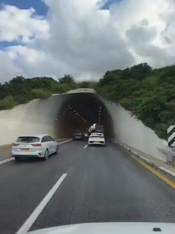 The video depicts a car traveling through a tunnel on a sunny day. The tunnel has a light-colored, arched ceiling with rows of lights. Several other cars are also traveling through the tunnel ahead. The road is paved with white lane markings. Vegetation is visible at the tunnel entrance. The sky is partly cloudy.