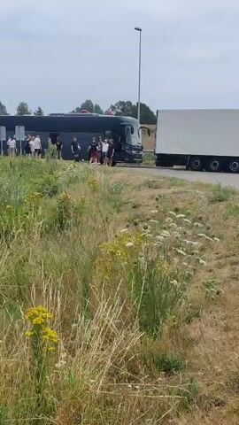 A black bus is parked on the side of a road, with a white truck parked next to it. Several people are standing around the bus, some appear to be getting off or on. The area around the bus is overgrown with tall green grass and some wildflowers. The sky is overcast.