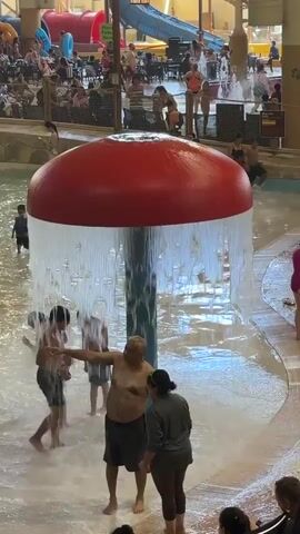 The video shows an indoor water park. In the foreground, a large, red, mushroom-shaped fountain sprays water onto a group of people. A man in dark shorts is standing directly under the spray. To his left, two younger males are also under the spray, one wearing dark trunks and the other wearing pink swim shorts. To his right, a woman in a dark shirt and dark pants watches. In the background, a lazy river with people floating on tubes can be seen, along with several water slides. Other people are sitting or standing around the pool area. The water park appears to be busy with many patrons.