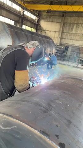 Two individuals are inside a large industrial building, likely a workshop or factory. The individual in the foreground, wearing a dark shirt, denim overalls, and welding gloves, is actively welding a large, curved metal object. Sparks are flying from the welding torch, illuminating the immediate area with a bright blue and orange glow. The welder is wearing a protective welding mask. In the background, another person is seated and also appears to be welding, with a similar bright light emanating from their work area. The environment appears industrial, with large metal structures, possibly tanks or pipes, in the background. The ceiling is high, with visible beams and lighting fixtures. The floor is concrete and appears somewhat worn. The overall scene depicts a manufacturing or fabrication process involving welding.