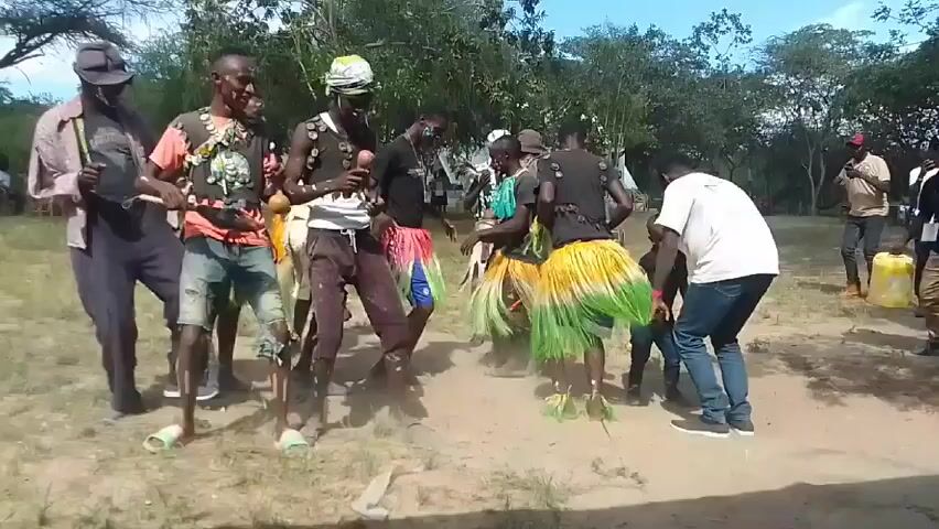 Here is a concise description of the video:

The video shows a group of approximately 8 adults and 2 children performing a folk dance in an outdoor setting. The dancers are in a dusty area with scattered trees and shrubs in the background.  Some of the dancers are wearing casual clothes, while others are adorned in traditional clothing, including beaded necklaces and colorful skirts. One dancer plays a maraca. At least two individuals are recording the dance on their phones. The overall impression is that of a cultural celebration or performance taking place.