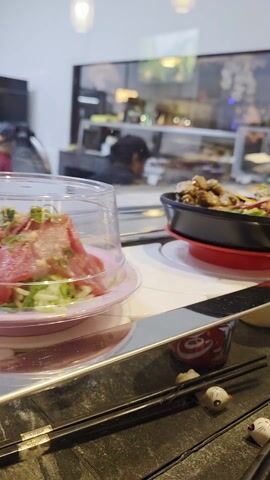 Here's a detailed description of the video:

The video shows a conveyer belt sushi restaurant with several dishes moving past the camera. The closest plate has a dome cover with what appears to be raw fish inside, on a bed of something green and white.  Next to it is a black dish, resembling a small frying pan, containing what looks like cooked beef or chicken with a salad. Further along, under a plastic cover, is a slice of cake. 

The conveyor belt itself is white with reflective metal accents.  In the foreground, on the table, are a pair of black chopsticks resting on ceramic cat-shaped chopstick rests.  A small red Coca-Cola themed container is visible in front of the chopsticks.  

The background shows the restaurant’s kitchen area behind a glass window, with people working inside. The environment is generally well-lit. There is some background noise indicative of the restaurant environment.