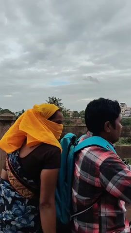 Two people are riding on a motorcycle. The person in the back is a woman wearing a dark brown shirt, a blue and black floral patterned saree, and a yellow headscarf that covers her face. She is wearing a blue backpack. The person in the front is a man wearing a red, black, and white plaid shirt and a blue backpack. They are on a road with a bridge railing and trees in the background, under a cloudy sky. The motorcycle is black.