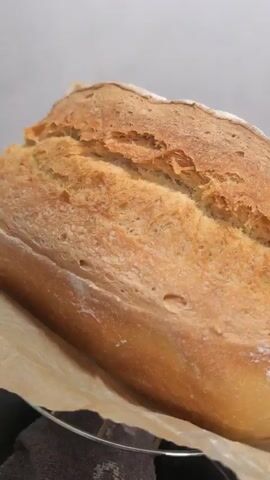 The video shows a close-up shot of a loaf of bread. The bread has a golden-brown crust with a deep score down the middle. It is dusted with flour and appears to be sitting on a piece of parchment paper. The background is blurred and out of focus, suggesting the focus is entirely on the bread. The lighting is soft and natural. In the final frames, a hand with pink nail polish is visible, reaching towards the bread.