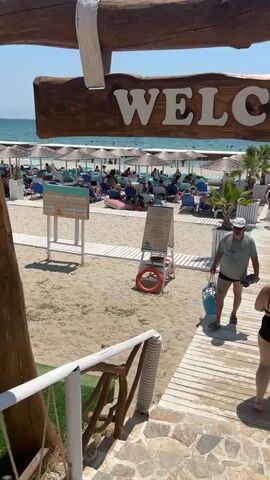 The video shows a beach scene with a "WELCOME" sign hanging from wooden beams. In the foreground, a wooden walkway leads down to the beach. Several people are walking on the walkway. One man is carrying a blue bag and wearing a hat and shorts. A woman in a swimsuit walks ahead of him. Further down the walkway, other people are heading towards the beach. The beach itself is covered with sand and has rows of lounge chairs and umbrellas. The sea is visible in the background with people swimming. To the right, there is a small hut with a thatched roof and a sign that says "CAMEO". A hanging chair with cushions is also visible, as is a tall speaker mounted on a wooden pole. The overall environment is a sunny beach resort.