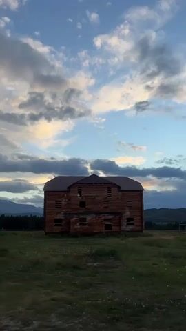 The video opens with a long shot of a large, unfinished wooden building with a dark roof, set in a grassy field under a cloudy sky. Mountains and a forest are visible in the background. The camera pans to the left, revealing a smaller, darker building with a porch. Several people are seated on chairs outside the smaller building, and a car is parked nearby. A red object, possibly a jacket, lies on the ground in front of the wooden building. The video continues to pan left, showing more of the smaller building and the surrounding landscape, with the unfinished wooden building remaining visible in the background.