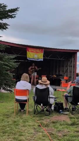 A wide shot shows an outdoor performance. A person is playing a guitar and singing on a raised wooden stage. A flag hangs behind the performer. Three people are seated in chairs facing the stage, and one person is clapping. The stage is under a simple red roof supported by wooden posts. There is grass in the foreground, and trees on the left side of the frame. The sky is cloudy.