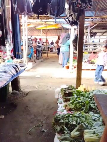 The video opens with a view from within an open-air market. In the foreground, on the right, is a stall displaying an abundance of fresh green vegetables. The ground appears to be dirt. To the left, a stall displays clothing, including what looks like trousers or jeans. In the middle ground, several people are visible. A person wearing a light blue top and light-colored trousers is walking away from the camera. Further down the market aisle, more people are milling about. A light blue motorcycle is parked. In the background, the market appears to extend further with stalls and people. The overall environment suggests a busy, typical market scene.