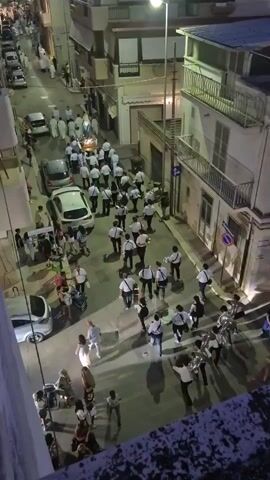 This is an aerial, medium-distance video recording of a religious procession on a narrow street at dusk. A group of people, many in white robes, are carrying a float with a statue on it. Behind them, a brass band in white shirts and dark pants marches, playing instruments. On either side of the street, people are watching the procession, some are standing, some are sitting in chairs. Several parked cars are visible on the street. The buildings on either side of the street are multi-story structures, with balconies and windows. The street is paved and appears to be in an urban or town setting. The lighting suggests it is late afternoon or early evening.