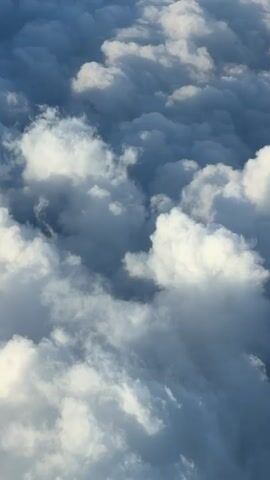 This video is an aerial view of clouds from above. The clouds are a mix of white and dark gray, suggesting varying levels of density and light. The camera appears to be moving slowly, offering a perspective as if from an airplane. The overall environment is atmospheric, with no discernable landforms or human presence. The lighting indicates it could be daytime, possibly during sunrise or sunset given the dramatic contrasts in cloud coloration.