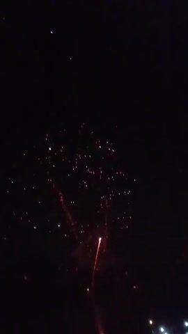 The video captures a fireworks display against a dark night sky. Several fireworks are visible, with various colors and patterns. One prominent firework in the upper right quadrant is a circular burst of orange and yellow sparks. Below it, a single firework streak of red and gold descends. The sky is filled with scattered sparks and smoke. In the bottom section of the video, a faint line of lights suggests a distant horizon or perhaps observers on the ground. The overall scene is one of a nighttime celebration marked by pyrotechnics.