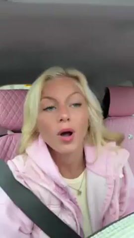 A young woman with fair skin and blonde hair sits in the driver's seat of a car. She is wearing a light pink puffer jacket over a light-colored shirt, and a seatbelt. She starts by making a kissing face, then smiles, then waves her hand to the music. The car interior features pink, quilted seat covers. Loud music is playing.