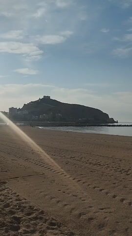 The video shows a panoramic view of a beach with a castle on a hill in the distance. The camera pans slowly from left to right, revealing the sandy shore, the calm ocean, and the blue sky with scattered clouds. The sun is shining, casting a warm light on the scene. There are no people or objects visible in the foreground, but there appear to be some structures on the hill in the distance, possibly part of the castle. The overall impression is one of tranquility and natural beauty.