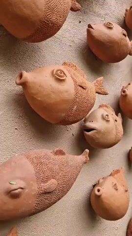 A close-up, panning shot shows a wall decorated with many clay fish sculptures. The fish are made of terracotta and vary in size and expression. Some have wide-open mouths, while others have small, circular openings. The sculptures are arranged in clusters and rows, creating a playful and whimsical display. The background is a textured wall, and the lighting is soft and natural, suggesting an outdoor setting.