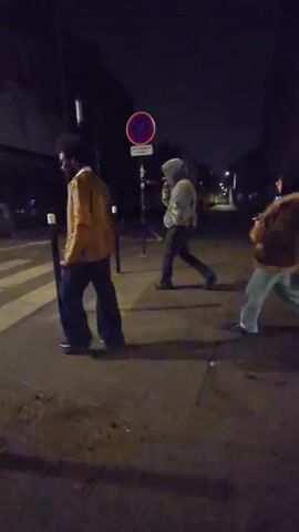 At night, three people are walking across a pedestrian crosswalk on a city street. The person closest to the camera, on the left, has dark hair and a beard and wears a brown jacket and dark pants. The person in the middle wears a hooded jacket and dark pants. The person on the right wears a brown jacket and light-colored pants. A “no parking” sign is visible at the edge of the crosswalk. The street is otherwise deserted.