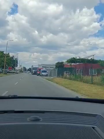 The video begins by showing the dashboard of a car. The camera then pans forward through the windshield, revealing a two-lane road with a white line dividing the lanes. Several vehicles are parked or driving on the right side of the road. These include a large white semi-truck, a red van, and a silver van. Further down the road, more vehicles can be seen, including cars and what appears to be another truck. The road is lined with buildings and greenery on both sides. The sky is mostly cloudy with patches of blue. The video focuses on the vehicles and the road ahead, giving the impression of a vehicle in motion. The semi-truck on the right side of the road has the word "delamode" and "International Logistics" written on its trailer in large blue letters.