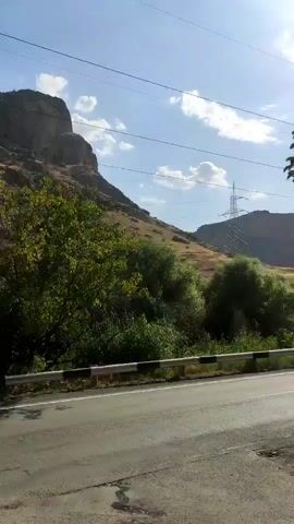 The video shows a mountain landscape. The terrain is rocky and barren in parts, with some vegetation, including trees and bushes, present on the slopes and in the foreground. A road is visible in the lower portion of the frame, with a guardrail separating it from the vegetation. Power lines stretch across the sky. The sky is blue with some clouds, indicating daylight. The camera appears to be moving, possibly from a vehicle.