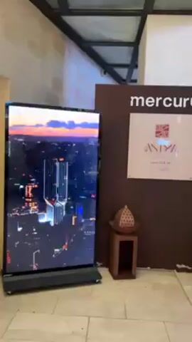 The video shows an interior hallway with a screen displaying images of a restaurant. A sign next to it says "mercury" with the word "ANTINA" and "Level 33 & 34".  There are two small wooden stools with decorative objects. The camera moves to show a modern building exterior with outdoor seating and lights. People are walking around, and there are trees and other greenery.