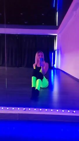 A blonde woman in a neon green and black athletic outfit sits on the floor in front of a large mirror in a dimly lit room with purple and blue lighting. She is holding a phone and appears to be taking a selfie. The floor is made of wood, and the walls are dark. There are blue LED lights along the bottom of the mirror.